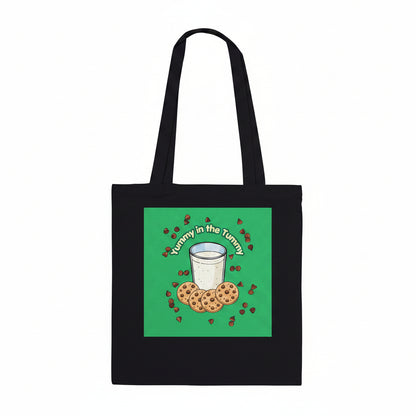 A black canvas tote bag with two shoulder straps, featuring a square green graphic print on the front. The graphic shows a white glass of milk surrounded by chocolate chip cookies and small red hearts, with the text 'Tummy in the Tummy' arched above them.