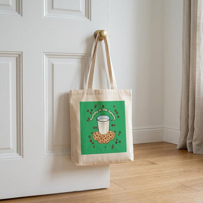 A beige canvas tote bag with a green square graphic hanging from a brass doorknob on a white paneled door. 