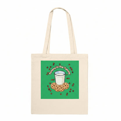 A natural canvas tote bag with a green square graphic on the front featuring an illustration of chocolate chip cookies, milk, and the text 'Yummy in the Tummy' in a retro curved font.